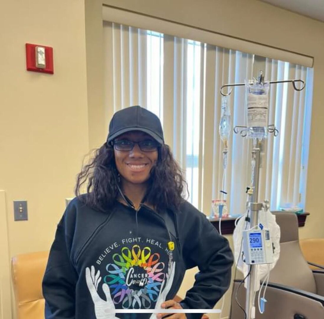 A smiling woman in a cap and colorful sweatshirt in a medical setting.